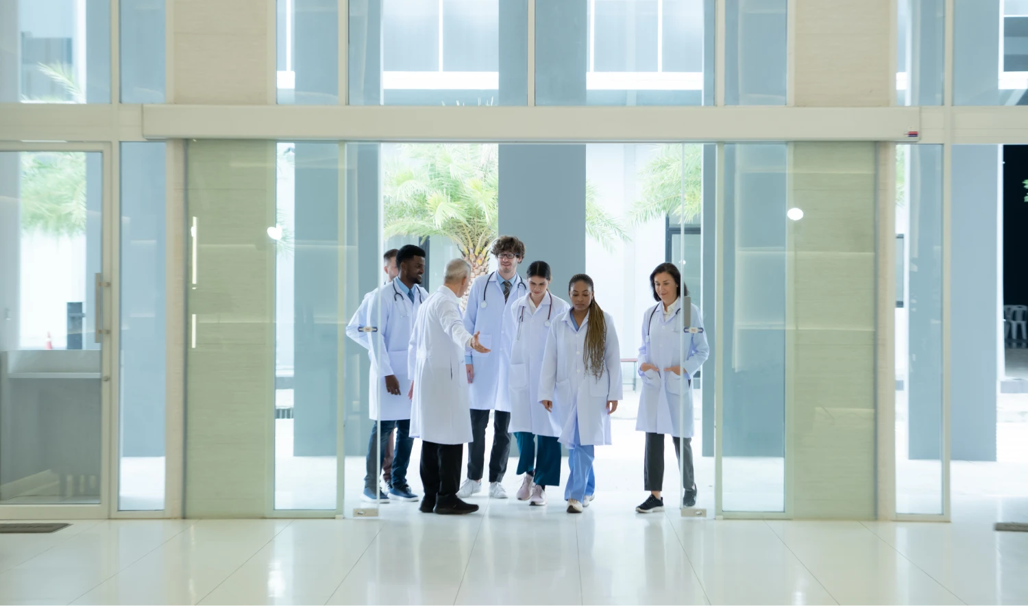 A group of medical professionals in white coats stand in a modern building hallway with glass doors.