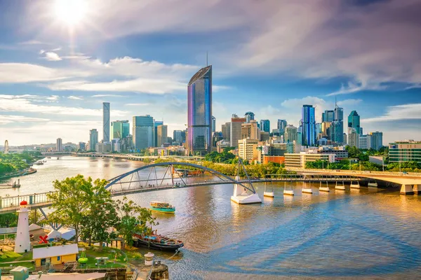 A river scene with a modern city skyline, featuring high-rise buildings and a prominent bridge under a sunny sky.