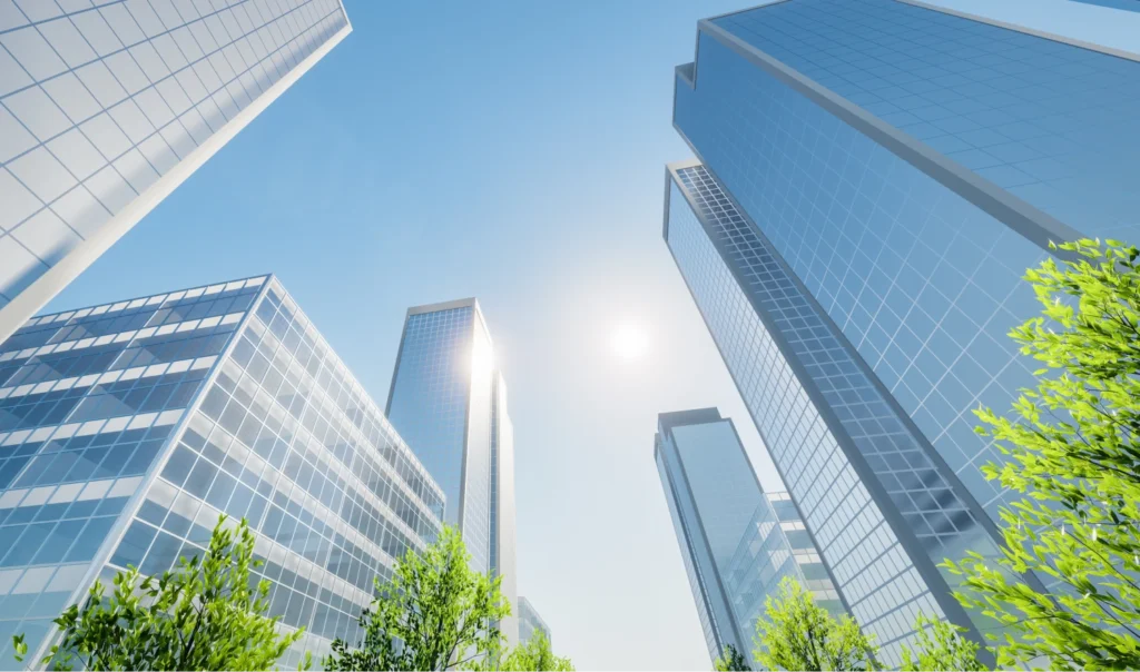 A city skyline with several tall glass skyscrapers under a clear blue sky and bright sun, surrounded by green trees.