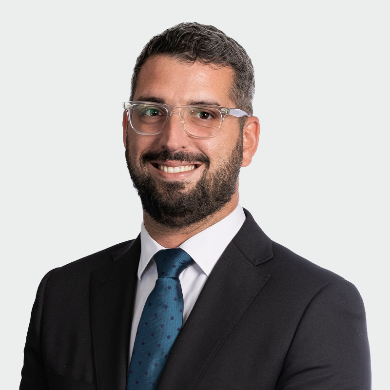 A man with glasses, a beard, and a mustache is smiling while wearing a suit and blue tie against a plain background.