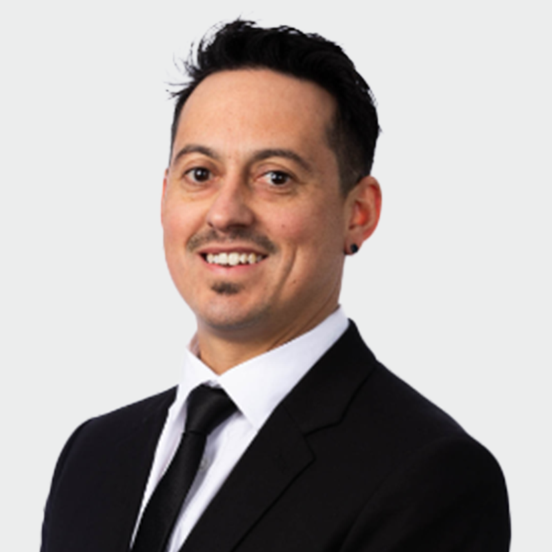 A man in a black suit, white shirt, and black tie is smiling against a plain white background.