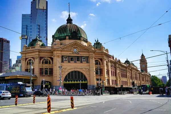 A bustling street scene in front of Flinders Street Railway Station in Melbourne, Australia, with trams, pedestrians, and surrounding skyscrapers under a clear blue sky.