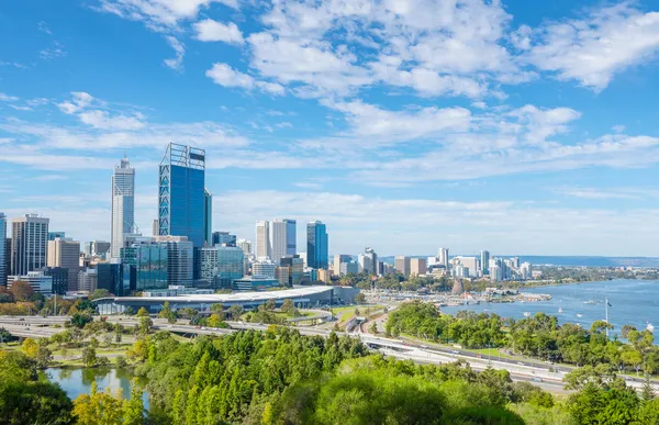 City skyline with tall buildings in the background, lush green park, and a river in the foreground under a partly cloudy sky.