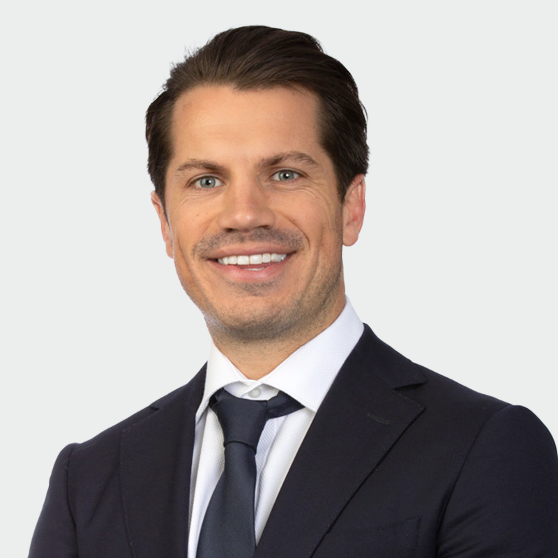 A man in a dark suit and navy tie smiles against a plain light background.